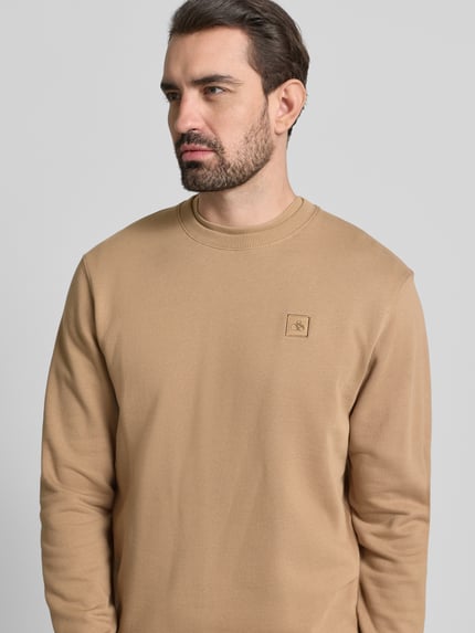 Männermodel draagt T-Shirt met Label-Stitching van Scotch & Soda, regular fit, ribkraag, pure katoen, neutrale beige kleur, bovenlichaam close-up.