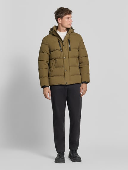 Männermodel trägt Steppjacke mit Kapuze von BRAX in Unifarben mit Logo-Patch und elastischen Abschlüssen, Ganzkörperansicht von vorne.
