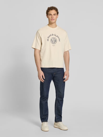 Männermodel trägt Relaxed Fit T-Shirt aus reiner Baumwolle Modell 'BOXYNORMAN GRAPHIC' von SELECTED HOMME in Beige mit Motiv-Print, Ganzkörperansicht von vorne.
