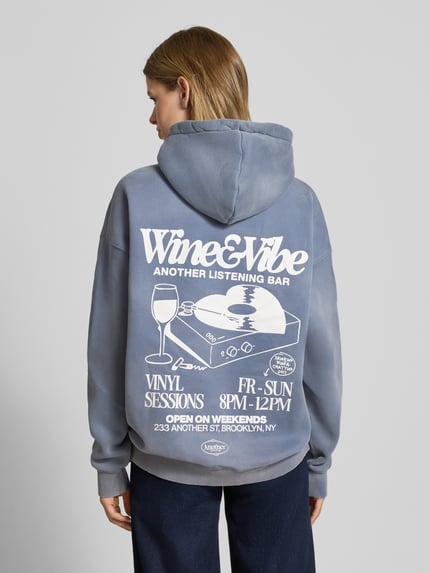 Kobiece model prezentuje Oversized Hoodie mit Känguru-Tasche Modell 'Wine & Vibe' od ANOTHER COTTON LAB, mieszanka bawełny, efekt znoszenia, nadruk z logo, kieszeń kangurka, widok całej sylwetki z tyłu.