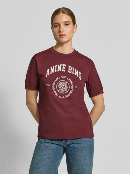 Kobiece modelka nosi damski t-shirt z nadrukiem z logo marki Anine Bing, wykonany z bawełny, prosty krój, okrągły dekolt, długość tyłu w rozmiarze S: 58 cm, widok całej sylwetki z przodu.