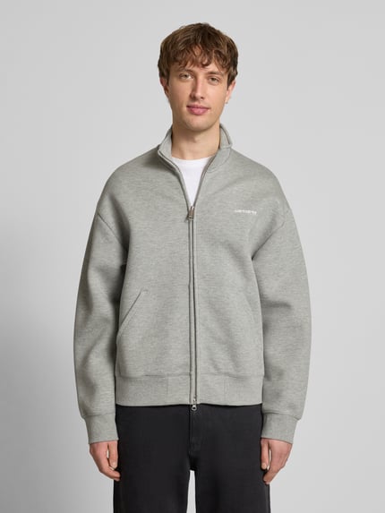 Męski model nosi Sweatjacke mit Stehkragen od Carhartt Work In Progress, bawełniany mix, luźny krój, stójka, zamek, naszywka z logo, kieszenie boczne, ściągacze, szary kolor, długość tyłu w rozmiarze M: 68 cm, długość rękawa w rozmiarze M: 61 cm, widok całej sylwetki z przodu.