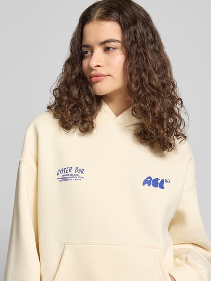 Frauenmodel trägt Hoodie mit Label-Print und Känguru-Tasche von ANOTHER COTTON LAB, cremefarbener Baumwoll-Mix, Relaxed-Fit, Nahaufnahme Oberkörper.