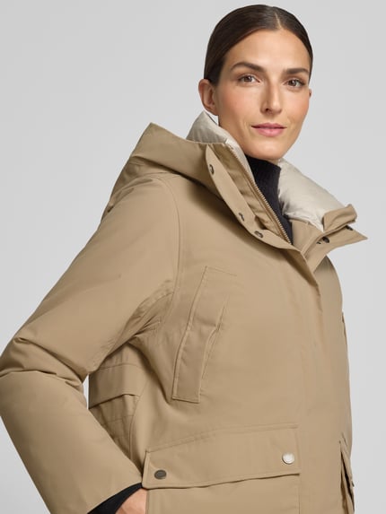 Frauenmodel trägt Regular Fit Parka mit Kapuze Modell 'Melody' von Didriksons in Beige mit aufgesetzten Taschen und elastischen Ärmelabschlüssen, Nahaufnahme Oberkörper.