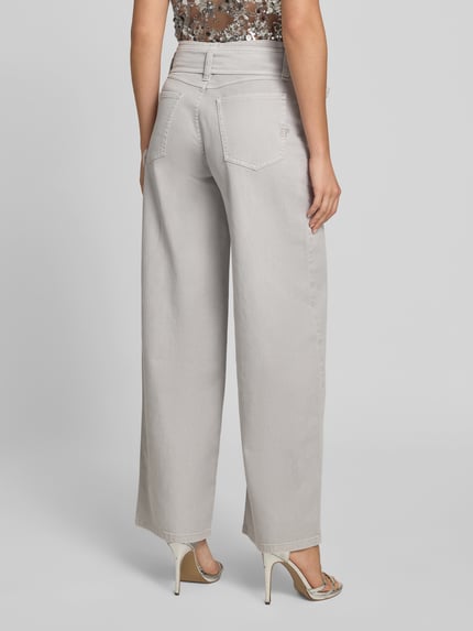 Frauenmodel trägt Wide Fit Jeans mit integriertem pasek od BAUM & PFERDGARTEN, mieszanka bawełny, krój wide fit, 5 kieszeni, jasnoszary kolor, długość wewnętrzna nogawki 82 cm, szerokość w rozmiarze 27: 38 cm, widoczny pasek, zapięcie na guzik i zamek błyskawiczny, widoczne faktury szwów, widok na całą sylwetkę z tyłu.