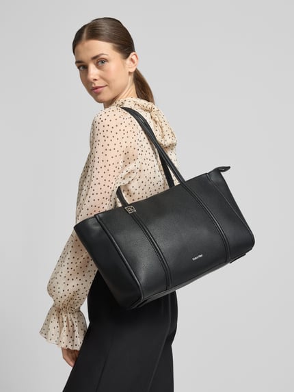 Vrouwenmodel draagt zwarte Shopper met Label-Detail van CK Calvin Klein met ritsvak, draaghengsels, schouderriem, hoofdvak, binnenvak, 47x26x16 cm, schouderriem 63 cm, polkadot blouse, geheellichaamsaanzicht achteraan.