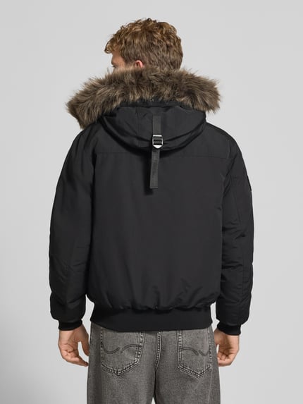 Männermodel trägt schwarze Bomberjacke mit Kunstfellkapuze, Label-Patch, Modell 'Everest' von Superdry, Rückenansicht, Ganzkörperansicht Rückansicht. Regular Fit, Reiß- und Druckknopfverschluss, Pattentaschen, Rückenlänge 71 cm, Schulterbreite 50 cm, Brustweite 65 cm, Ärmellänge 70 cm.