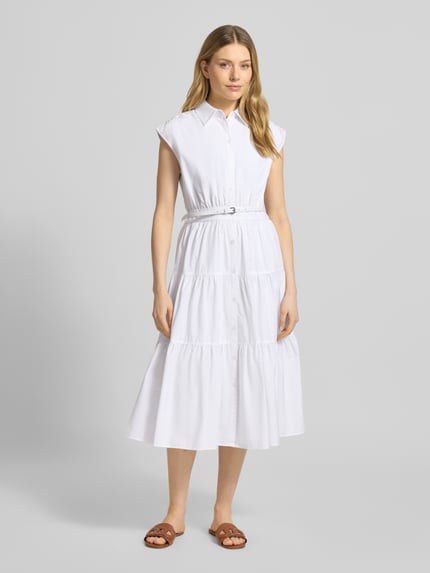Frauenmodel trägt Hemdblusenkleid mit Gürtel und Knopfleiste von MICHAEL Michael Kors aus weißer Baumwolle mit Volantsaum, Ganzkörperansicht von vorne.