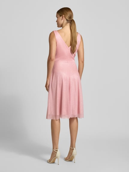 Frauenmodel trägt Knielanges Cocktailkleid mit V-Ausschnitt von TROYDEN COLLECTION in Rosa, Chiffon, tailliert, knielang, breite Träger, Rückseitiger V-Ausschnitt, Reißverschluss, feine Textur, dezenter Glanz, Ganzkörperansicht Rückansicht.