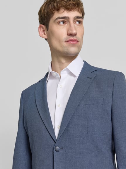 Mannelijk model draagt Slim Fit Sakko met borstleistentasche van Selected in gemêleerde look met fijn geweven motief, reverskraag, eenrijige 2-knoopsluiting, knopen, paspelzak op de borst, strookzakken en binnenzak, detailopname bovenlichaam.