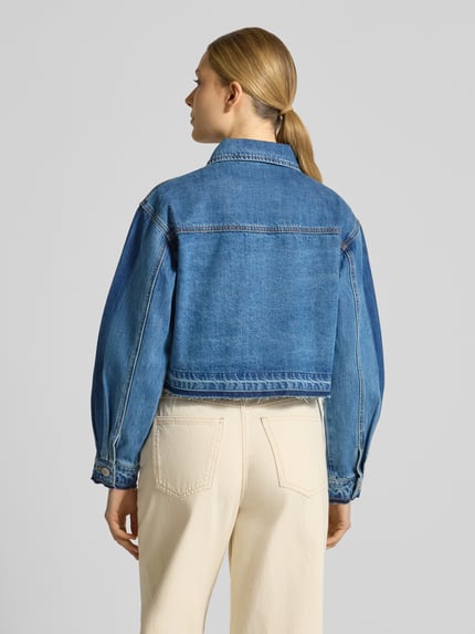Modelka nosi kurtkę jeansową z dodatkiem wiskozy model 'DENZEL' od SUNCOO PARIS, mieszanka materiałów, prosty krój, wykładany kołnierz, zamek błyskawiczny, szerokie ramiona, długość tyłu w rozmiarze S: 47 cm, szerokość ramion w rozmiarze S: 48 cm, szerokość klatki piersiowej w rozmiarze S: 53 cm, długość rękawów w rozmiarze S: 56 cm, niebieski denim, gładka faktura, całkowity widok z tyłu.