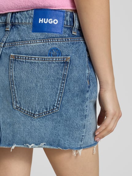 Vrouwenmodel draagt Mini-jeansrok uit puur katoen model 'GALANIE_1_B' van Hugo Blue, denimlook met studs, labelpatch, 5-pocketmodel, contrastnaad, knoopsluiting, open zoom, smal, minilengte, detailopname achterzijde.