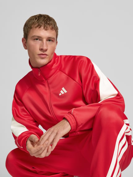 Männermodel trägt Trainingsjacke mit Stehkragen und Logo von adidas Sportswear in Rot mit weißen Details, Material-Mix, Nahaufnahme Oberkörper.