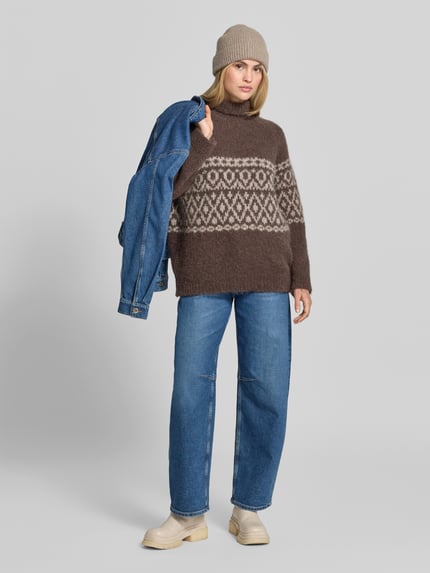 Frauenmodel trägt Pullover aus Alpaka mit Rollkragen Modell 'CRISTINE' von BAUM & PFERDGARTEN in brauner Strickoptik mit geometrischem Jacquardmuster, strukturierte Textur, lange Raglanärmel mit gerippten Abschlüssen, kombiniert mit blauer Jeans und beiger Mütze, Ganzkörperansicht von vorne.