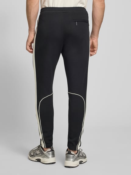 Männermodel trägt Slim Fit Tracksuit mit Logo-Stitching Modell 'Egeo' von SERGIO TACCHINI, czarny kolor, elastyczny bawełniany materiał, spodnie z białymi lamówkami, detal z logo, krój slim fit, Szerokość w rozmiarze M: 42 cm, długość tyłu: 64 cm, długość nogawki wewnętrznej: 73 cm, widok całej sylwetki z tyłu.