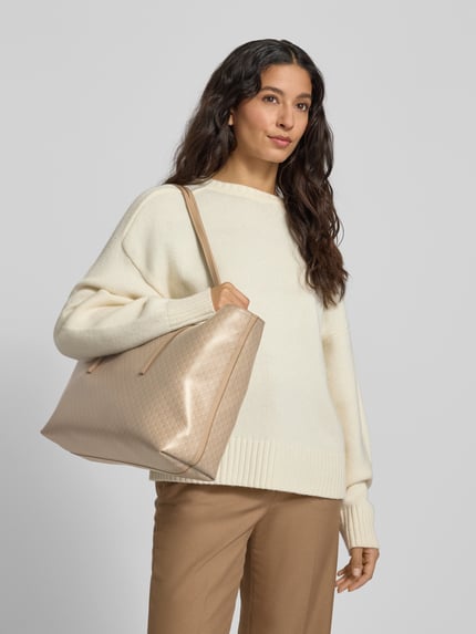 Vrouwenmodel draagt Tote Bag met Allover-Logo-Print van CK Calvin Klein in lichtbeige, open hoofvak en smalle draagband, Ganzkörperansicht van voren. Afmetingen: breedte 40 cm, lengte 28 cm, diepte 13 cm, schouderriemlengte 68 cm.