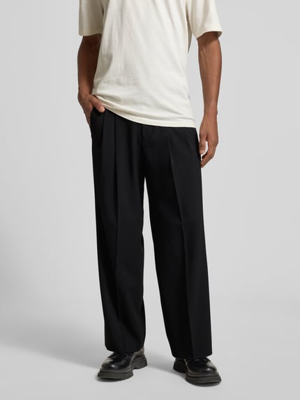 Männermodel trägt Wide Fit Hose mit Bügelfalten Modell 'Leek' von Drykorn, schwarzer Material-Mix mit Viskose-Anteil, Ganzkörperansicht von vorne.