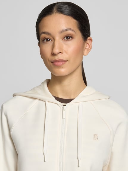 Frauenmodel trägt Regular Fit Sweatjacke aus Viskose-Mix mit Logo-Stitching von ARMANI EXCHANGE in Creme, Nahaufnahme Oberkörper. Elastischer Viskose-Mix, Logo-Stitching, Kapuze mit Tunnelzug, Reißverschluss, elastische Abschlüsse.