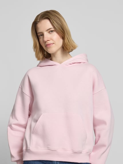 Vrouwenmodel draagt Regular Fit Hoodie met kangoeroezak model 'Iconic' van Gina Tricot, katoenmix, elastische boorden, lichtroze kleur, vooraanzicht bovenlichaam.