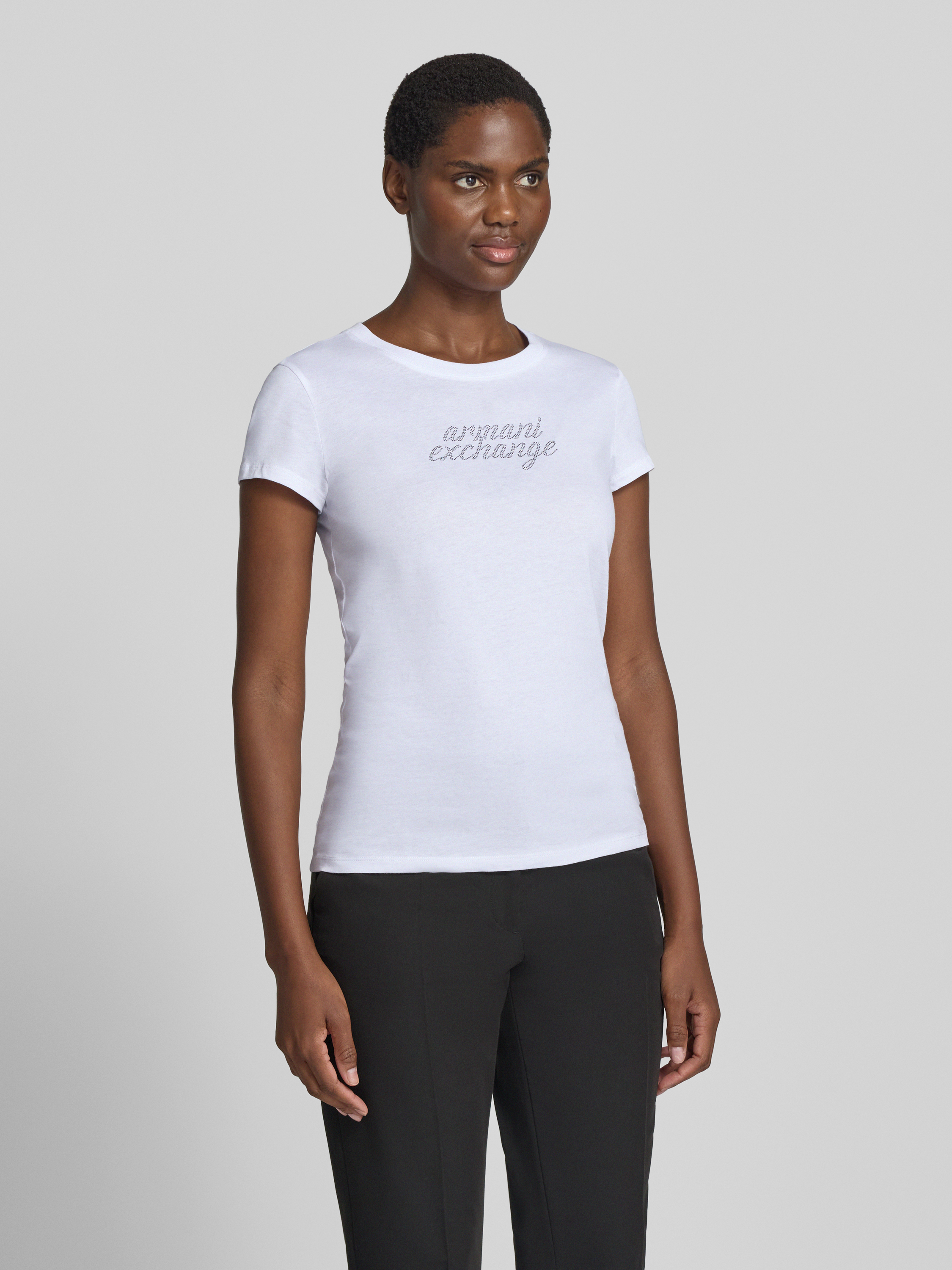 Frauenmodel trägt T-Shirt mit Ziersteinbesatz und Rundhalsausschnitt von Armani Exchange aus reiner Baumwolle, Slim Fit, Logo in Silber auf weißem Stoff, glatte Textur, Damen T-Shirt, Ganzkörperansicht von vorne.