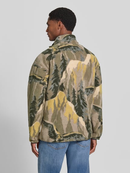 Männermodel trägt sweter z polaru Fleecepullover mit Label-Patch Modell 'HELVETIA' von Columbia w odcieniach zieleni, beżu i żółci, z motywem krajobrazu, prosty krój, stójka, długość tyłu w rozmiarze M: 73 cm, długość rękawów w rozmiarze M: 60 cm, Ganzkörperansicht Rückansicht