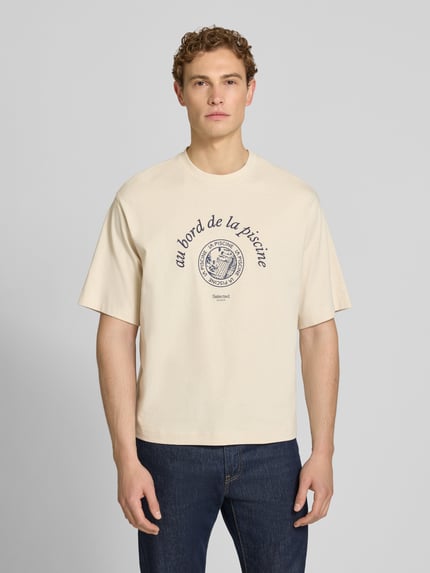 Männermodel trägt Relaxed Fit T-Shirt aus reiner Baumwolle Modell 'BOXYNORMAN GRAPHIC' von SELECTED HOMME in Beige mit Motiv-Print, Ganzkörperansicht von vorne.
