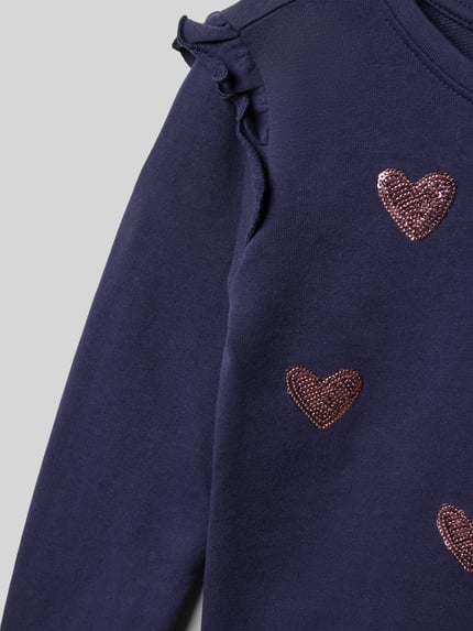 Mädchen trägt Sweatshirt mit Rüschen und Rundhalsausschnitt von Blue Seven, Reine Baumwolle, Regular-Fit, Motiv, Nahaufnahme Oberkörper.