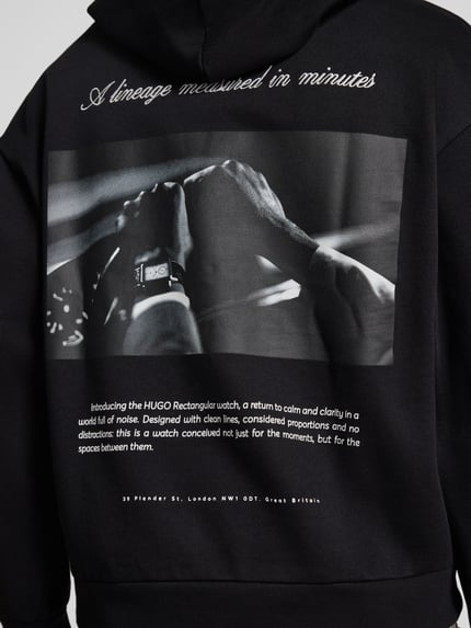 Mężczyzna nosi Oversized Sweatshirt aus reiner Baumwolle Modell 'Devrohood' od HUGO w kolorze czarnym z nadrukiem i teksturą dzianiny, Ganzkörperansicht Rückansicht. Kontext: bluza męska od HUGO.