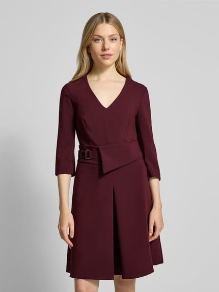 Frauenmodel trägt knielanges Cocktailkleid mit V-Ausschnitt von Rinascimento aus Material-Mix mit Viskose-Anteil, taillierter Schnitt, Taillengürtel, 3/4-Ärmel, Nahtreißverschluss, bordeauxrot, glatte Textur, Ganzkörperansicht von vorne.