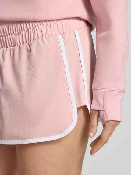 Frauenmodel trägt Running Shorts mit elastischem Bund von VARLEY in Rosa mit weißer Paspel, Material-Mix mit Stretch, Nahaufnahme Oberschenkel.