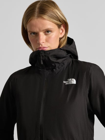 Frauenmodel trägt Jacke mit Kapuze und Zweiwege-Reißverschluss Modell 'HIKESTELLER' von The North Face in Schwarz, Nahaufnahme Oberkörper. Rückenlänge S 86 cm, Schulterbreite S 41 cm, Brustweite S 52 cm, Ärmellänge S 65 cm, Logo sichtbar, glatte Textur.
