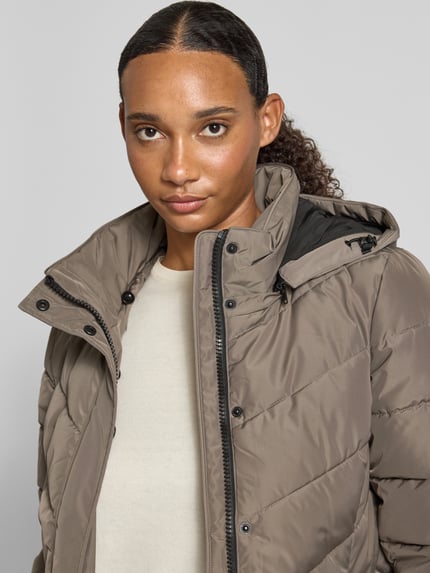 Frauenmodel trägt Regular Fit Jacke in Stepp-Optik von NAUTGÅRD mit Kunstfellbesatz und Eingrifftaschen, warme Beige- und Brauntöne, Nahaufnahme Oberkörper. Rückenlänge 68 cm, Schulterbreite 43 cm, Brustweite 47 cm, Ärmellänge 66 cm.