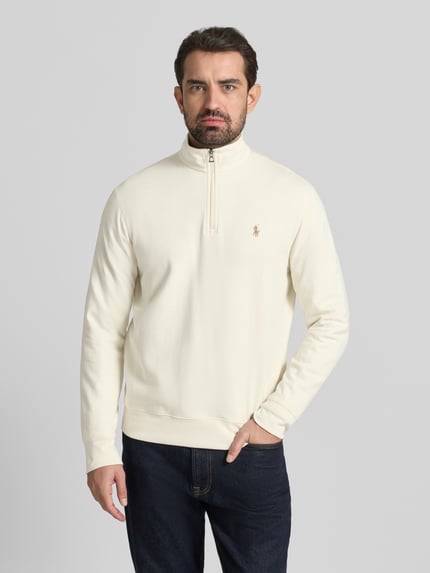 Męski model nosi Sweatshirt mit Label-Stitching od Polo Ralph Lauren w kolorze kremowym, bawełna z domieszką wiskozy, luźny krój, stójka, krótki zamek, prążkowane wykończenia, widok całej sylwetki z przodu.
