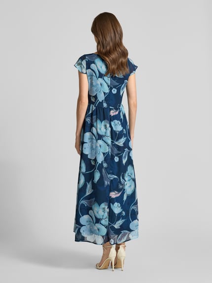 Modelka nosi długą sukienkę 'Maxikleid mit Schlüsselloch-Ausschnitt und Bindegürtel' od More & More w granatowe i jasnoniebieskie kwiaty, taliowany fason, okrągły dekolt, wycięcie w kształcie łezki, wiązany pasek, zamek błyskawiczny z tyłu, długość tyłu w rozmiarze 36: 135 cm, szerokość w rozmiarze 36: 48 cm, długość rękawów: 4 cm, pełny widok z tyłu.