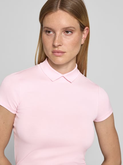 Modelka kobieca nosi Slim Fit T-Shirt mit hochschließendem Polokragen von Marc Cain z mieszanki bawełny i elastanu, krój slim fit, kołnierzyk polo, rękaw o dł. 1/2, jasnoróżowy kolor, gładka faktura, damski t-shirt, długość tyłu w rozmiarze 36: 59 cm, zbliżenie na górną część ciała.