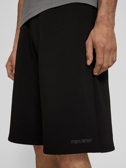 Männermodel trägt Sweatshorts mit Logo-Print von REVIEW in Schwarz, aus Material-Mix mit Baumwoll-Anteil, Loose-Fit, Nahaufnahme Oberschenkel.