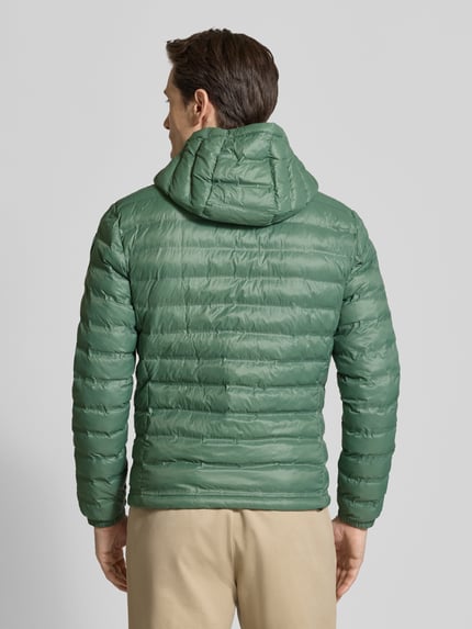 Męski model nosi Kurtka pikowana z zamkiem błyskawicznym dwustronnym model 'Charles' od Blauer Usa, krój regular fit, z kapturem, detal z logo, długość tyłu w rozmiarze M 65 cm, szerokość ramion w rozmiarze M 47 cm, szerokość w rozmiarze M 55 cm, długość rękawów w rozmiarze M 65 cm, zielona, matowa tekstura, prostokątne przeszycia, widok całej sylwetki od tyłu.