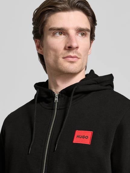 Männermodel trägt Sweatjacke aus Baumwolle Modell 'Daple212' von HUGO in Schwarz mit Kapuze, Label-Patch und Reißverschluss, Nahaufnahme Oberkörper.