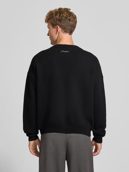 Männermodel trägt Relaxed Fit Strickpullover aus reiner Baumwolle von PROHIBITED, schwarze Farbe, lockere Passform, Label-Stitching, gerippte Abschlüsse, Ganzkörperansicht Rückansicht.