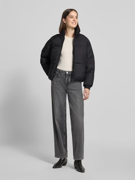 Frauenmodel trägt schwarze Pufferjacke mit Stehkragen Modell 'VMBRENDA' von VERO MODA, Cropped-Schnitt, Webware, seitliche Taschen, Ganzkörperansicht von vorne.