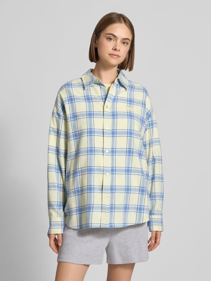 Frauenmodel trägt Relaxed Fit Hemdbluse aus reiner Baumwolle Modell 'Vivian' von OH APRIL, kariert in Blau und Gelb, Logo-Patch, Ganzkörperansicht von vorne.