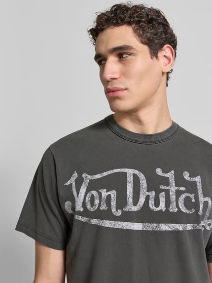 Männermodel trägt Relaxed Fit T-Shirt aus reiner Baumwolle von ANNARR in donkergrijs met labelprint, ronde hals, korte mouwen, nahaufnahme oberkörper