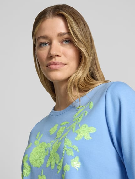 Kobiece model prezentuje T-Shirt aus Viskose-Mix mit Paillettenbesatz od Milano Italy, niebieski kolor, krój straight fit, okrągły dekolt, nadruk z motywem i cekinami, mieszanka wiskozy z dodatkiem stretchu, długość pleców w rozmiarze S: 57 cm, zbliżenie na górną część ciała.