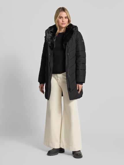 Frauenmodel trägt schwarze Jacke mit abnehmbarer Kapuze von NAUTGÅRD, seitliche Reißverschlusstaschen, Ganzkörperansicht von vorne.