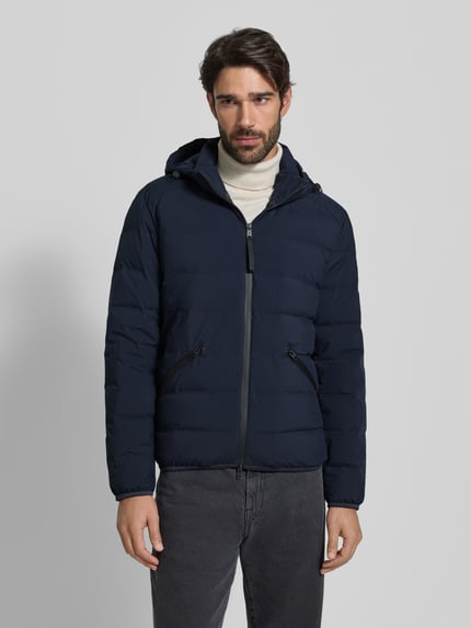Męski model nosi Regular Fit Daunenjacke mit Kapuze od Marc O'Polo, granatowa, jednokolorowa, kieszenie boczne, wodoszczelna, wiatroszczelna, oddychająca, wypełnienie z puchu i pierza, zamek błyskawiczny dwustronny, elastyczne lamówki; całkowity widok z przodu.