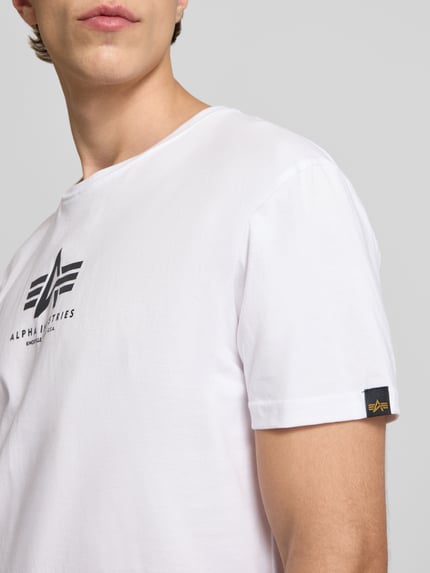 Männermodel trägt T-Shirt mit Logo und Rundhalsausschnitt van Alpha Industries in wit, katoen, regular fit, logo op borst, detailopname bovenlichaam.