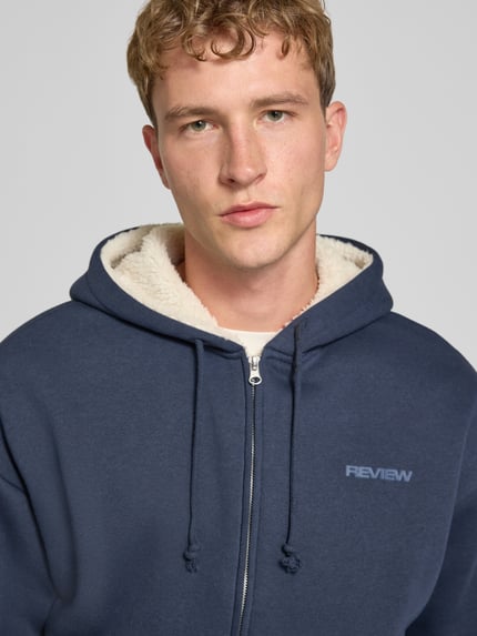 Männermodel trägt Regular Fit Sweatjacke aus Baumwoll-Mix mit Teddyfutter von REVIEW, dunkelblau, Logo-Detail, Nahaufnahme Oberkörper.