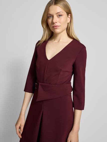 Frauenmodel trägt Knielanges Cocktailkleid mit V-Ausschnitt von Rinascimento in Dunkelrot, Material-Mix mit Viskose-Anteil, taillierter Schnitt, Taillengürtel, 3/4-Ärmel, Nahtreißverschluss, Nahaufnahme Oberkörper.