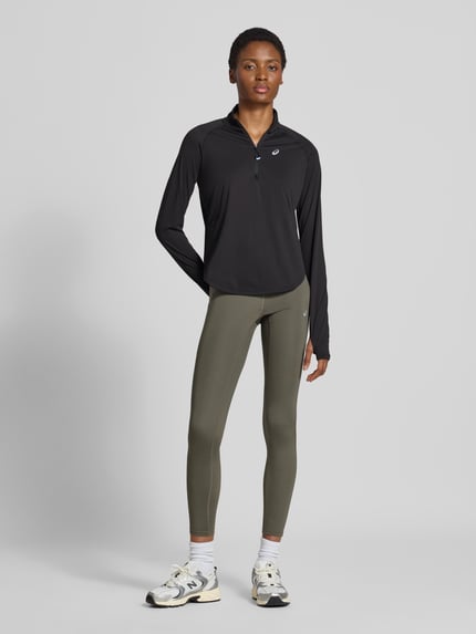 Frauenmodel trägt Longsleeve mit Troyer-Kragen Modell 'NAGINO' von ASICS TIGER in Schwarz, reflektierendes Logo, Regular Fit, Ganzkörperansicht von vorne.