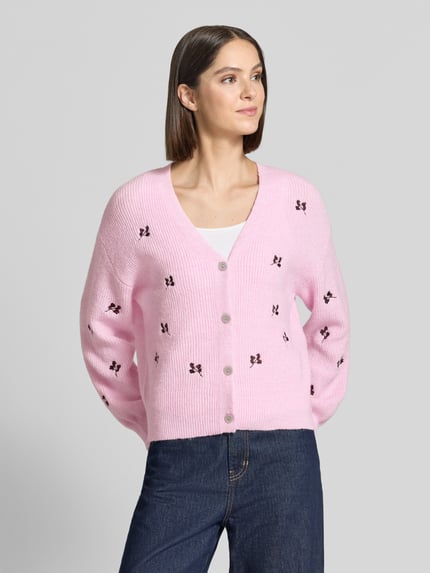Modelka kobieca ma na sobie Relaxed Fit Strickjacke mit Woll-Anteil Modell 'Fleur' od YAS, różowy kolor, kwiatowy haft, długi rękaw, guziki, widoczna tekstura dzianiny, długość pleców 58 cm, długość rękawa 54 cm, mieszanka materiałów z dodatkiem wełny, widok na górną część ciała z przodu.